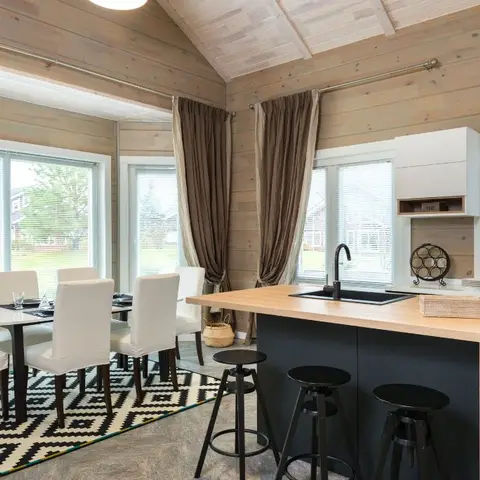 Log-cabin great room with a wood-panelled vaulted ceiling, a wood-top kitchen island with bar stools, and a slipcovered dining table on a graphic rug