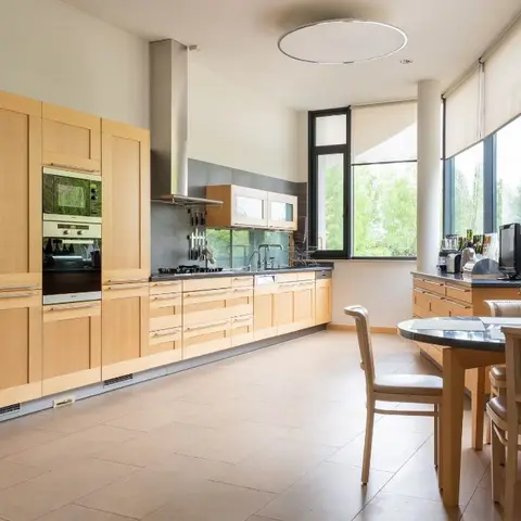 Long galley kitchen in light maple shaker cabinetry with a built-in oven and stainless chimney, grey tile backsplash