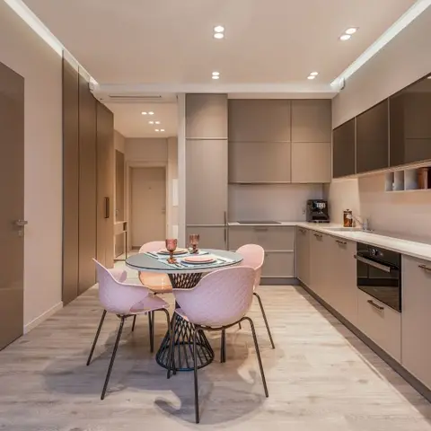 Long galley kitchen in matte taupe handleless cabinetry with a built-in oven, adjoining a small round dining table with quilted pink chairs.