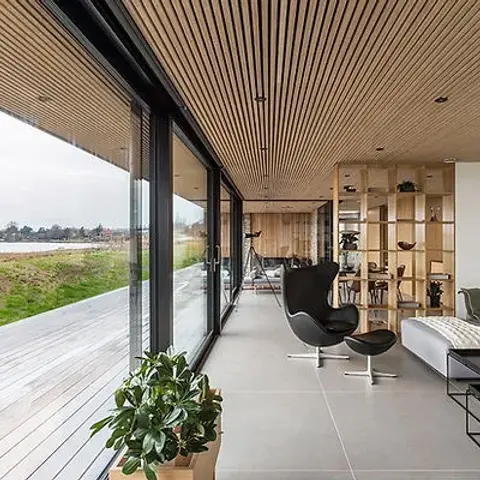 Long living room under a continuous oak-slat ceiling that runs out over the deck, with a black leather Egg chair and footstool facing a full-glass wall onto an