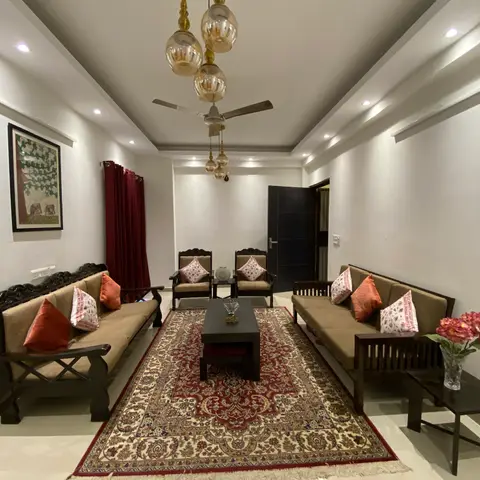 Long living room with facing carved-wood sofas around a red medallion Persian-style rug, a triple amber-glass cluster pendant and maroon curtains.