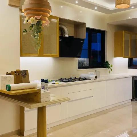 Long open kitchen shot at night: white handleless base units under a cream stone counter, cane-fronted wall cabinets in teak frames