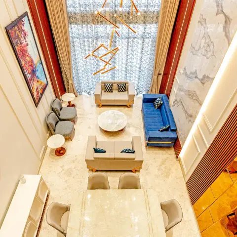 Looking down from a mezzanine onto a double-height living room in beige marble, where a royal-blue velvet sofa faces two cream loveseats around a round marble