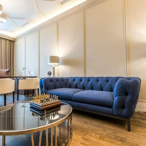 Lounge with a navy blue tufted chesterfield sofa against cream wall panelling, a faceted cove-lit ceiling, and a round mirrored coffee table with a chess