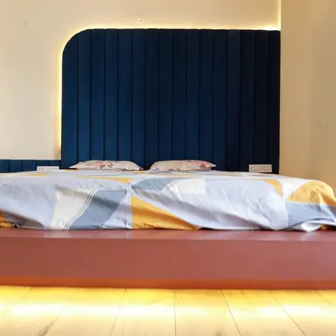 Low-angle bedroom shot where a navy fluted upholstered headboard is backlit against cream wall and the rust-red platform bed base floats on a strip of