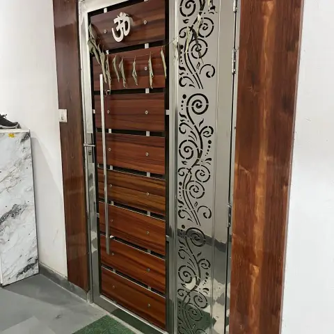 Main entrance safety door with wood-panel shutter, an Om motif, and a stainless-steel jaali side panel with floral scrollwork, framed in dark wood cladding.