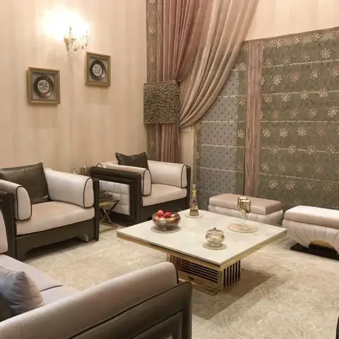 Majlis-style living room with dark-trimmed cream leather armchairs, a marble-and-gold coffee table, a richly patterned floral tapestry wall panel