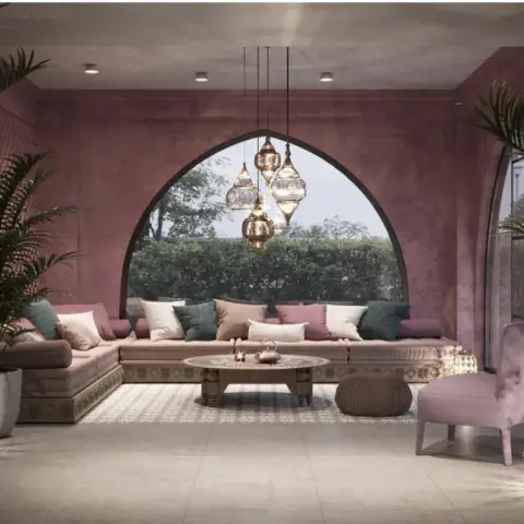 Majlis-style lounge in rose lime-plaster walls with a keyhole-arched window over a long low banquette piled with teal, mauve and blush bolsters
