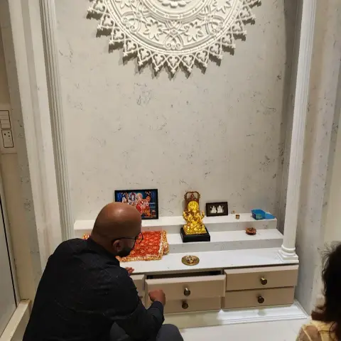 Marble-clad pooja niche with a carved white Lakshmi medallion between fluted pillars, a family at prayer before the drawer-fronted counter.
