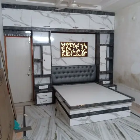 Marble-finish laminate platform bed with a backlit floral jaali headboard panel and matching overhead loft units, under a cove-lit false ceiling.