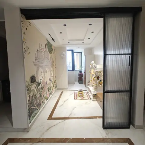 Marble pooja room entered through a black-framed fluted-glass sliding partition, with a Pichwai-style temple-and-peacock mural on the side wall and a carved