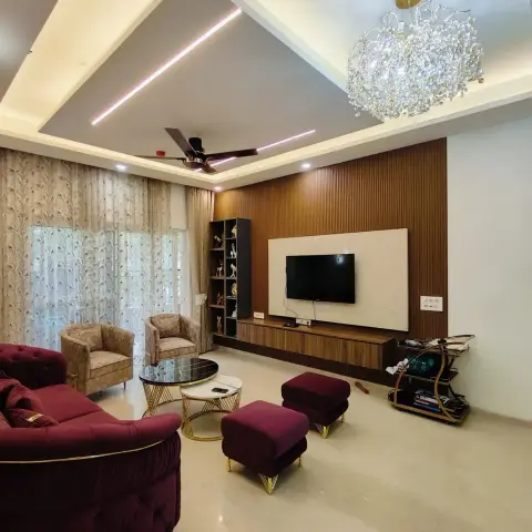 Maroon tufted chesterfield sofa and matching gold-legged ottomans face a fluted wood-panelled TV wall under a crystal chandelier and pink-lit cove ceiling.