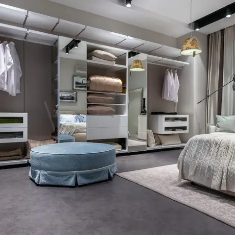 Master bedroom opening into a walk-in wardrobe with mirrored panels and open shelving, styled with a round blue tufted ottoman, pendant lamps