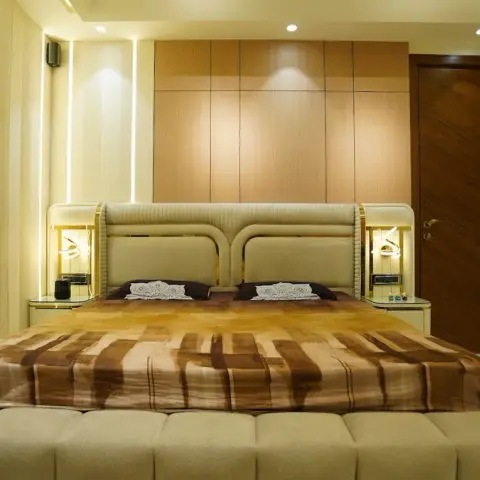 Master bedroom shot straight down the bed to a teak-veneer panel grid headboard wall, a curved cream headboard with brass trim between lit bedside niches