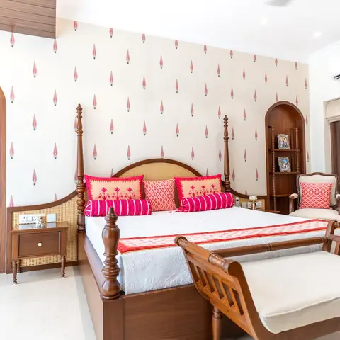 Master bedroom with a carved wood four-poster bed and cane headboard against a block-print pink wallpaper wall, with an arched wooden display alcove.