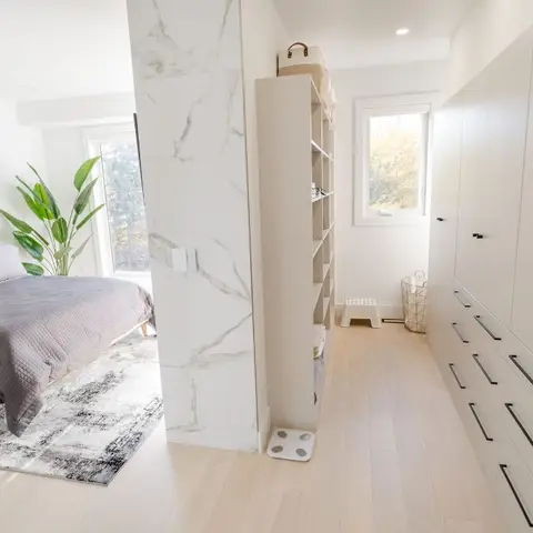 Master bedroom with a grey upholstered bed and abstract rug, opening through a marble-clad column into a walk-in wardrobe with floor-to-ceiling white cabinetry.