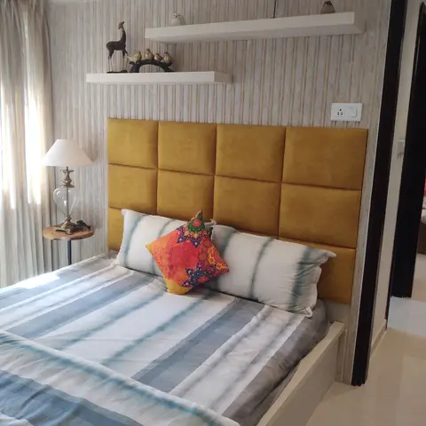 Master bedroom with a mustard-yellow tufted panel headboard against striped wallpaper, twin floating shelves styled with brass and wood curios