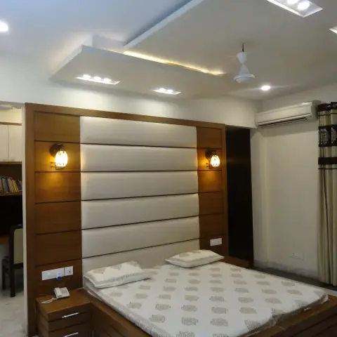 Master bedroom with a wood-and-cream padded headboard wall lit by sconces, a boxed cove-lit false ceiling, and a built-in study nook to the side.