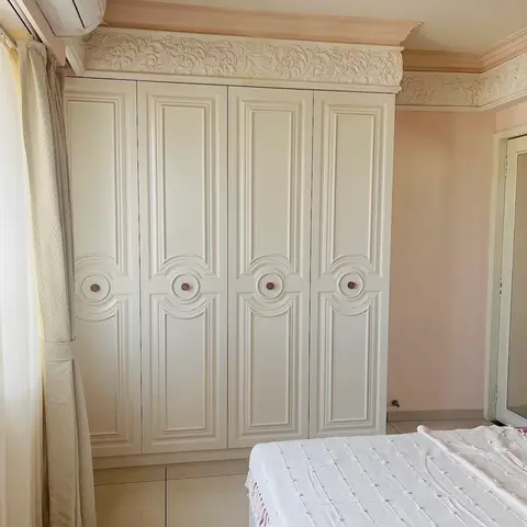 Master bedroom with an ornate ivory carved wardrobe under a floral cornice, set against blush pink walls.