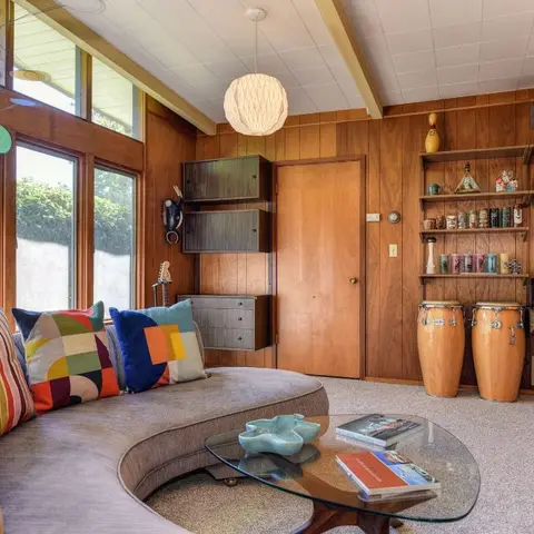 Mid-century living room with wood-panelled walls, a curved grey sofa styled with colour-block cushions, a glass kidney-shaped coffee table
