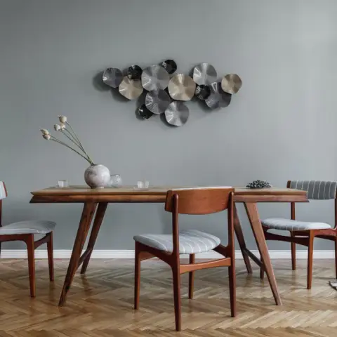 Mid-century teak dining table and striped-seat chairs on herringbone parquet, under a metal wall sculpture of overlapping brushed discs on a grey-green wall.