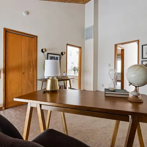 Mid-century trestle writing desk with a globe and books, set against a white wall with a small gallery of framed art and a wood-panelled door.
