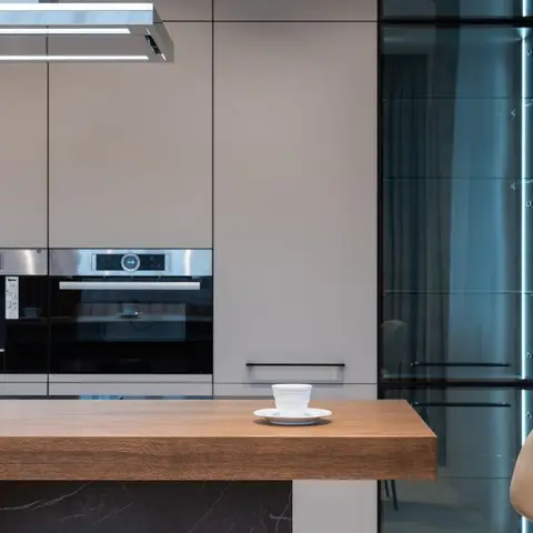 Minimal grey kitchen with built-in wall ovens, a coffee machine niche and an illuminated glass display cabinet beside a wood breakfast counter.