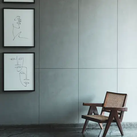 Minimalist corner with a caned wood armchair against a grey stone-panel wall hung with a trio of line-art portraits.