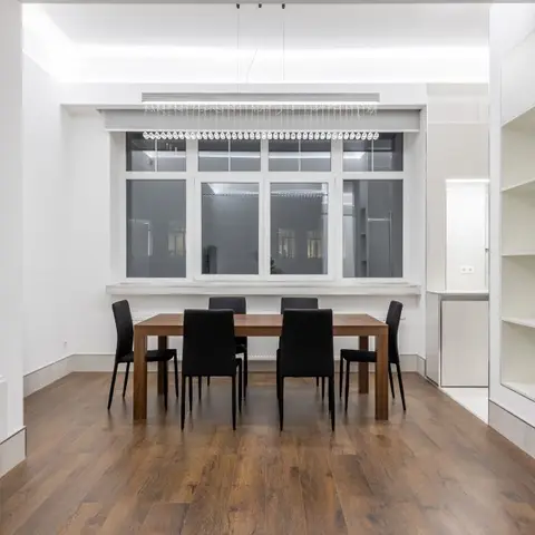 Minimalist dining nook with a walnut table and black upholstered chairs beneath a linear crystal-drop pendant, wood flooring and a large crittall-style window
