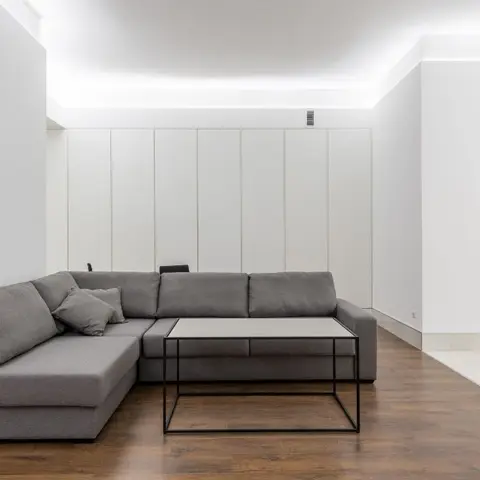 Minimalist living room with a grey L-shaped sectional facing a black-frame glass coffee table against a tall vertical-panelled wall.