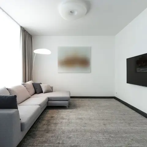 Minimalist living room with a grey L-shaped sectional sofa, a wall-mounted TV on a plain white wall, and a soft-focus abstract art print beside a floor lamp.