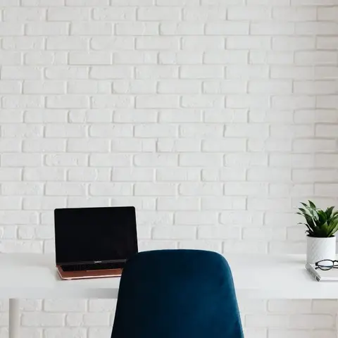 Minimalist study nook with a white desk, navy velvet chair, and a laptop set against a whitewashed brick wall