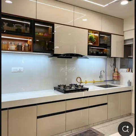 Modular kitchen with glossy beige shutters, a lit glass-front display cabinet showing jars and crockery, white quartz counter, and a ceiling cove LED strip.