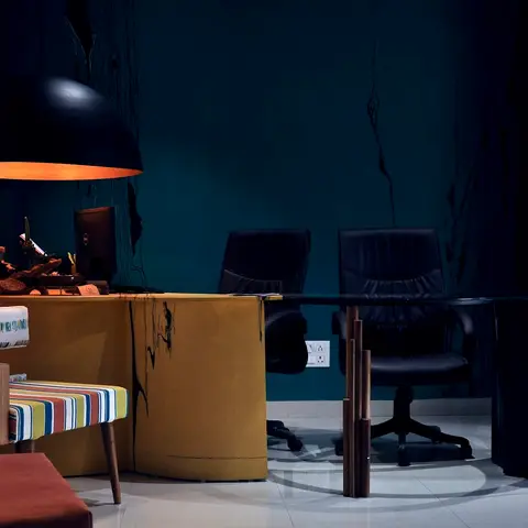 Moody home study with a curved mustard-yellow desk and black leather chairs against a dark teal textured wall, lit by an oversized black dome pendant