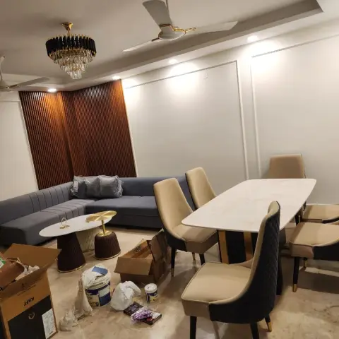 Moving-in day in an open living-dining: a grey channel-tufted L sofa still in its plastic wrap sits against a fluted teak panel wall under a black-and-crystal