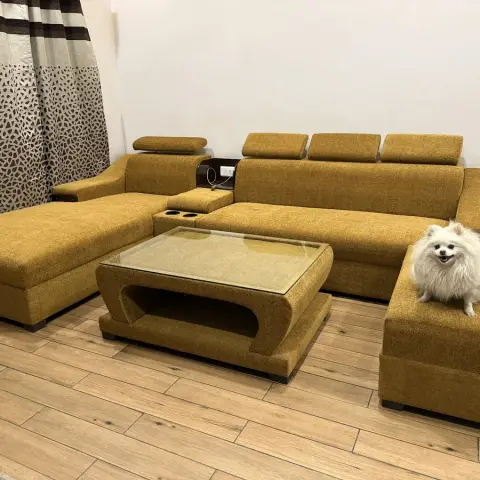 Mustard-yellow L-shaped fabric sectional sofa with adjustable headrests and a glass-top coffee table on wood-look tile flooring.