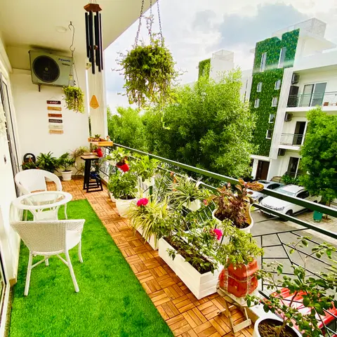 Narrow apartment balcony with white wicker chairs on artificial turf beside wood-tile decking, lined with potted flowering plants and a hanging metal wind chime