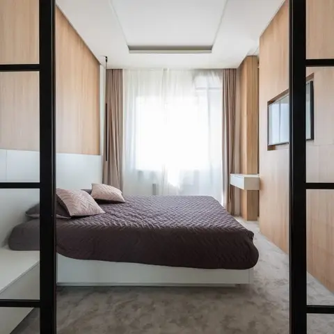 Narrow bedroom seen through black-framed glass partition doors: oak-veneer walls, a brown quilted bed on a white plinth and a TV recessed into the wood