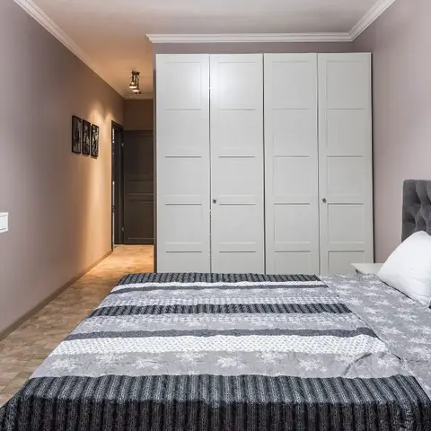Narrow bedroom with a grey button-tufted headboard and striped grey bedding, a white four-door panelled wardrobe filling the end wall