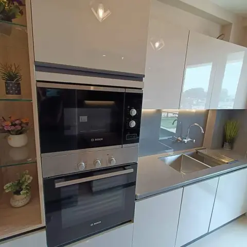Narrow white high-gloss galley kitchen with a stacked Bosch built-in microwave and oven tower beside a grey lacquered-glass backsplash and undermount sink