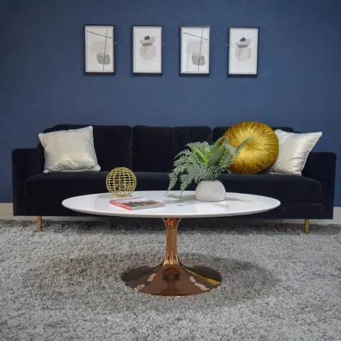Navy velvet sofa on brass legs under four small abstract prints on a deep blue wall, with a white oval tulip table on a copper base standing on a grey shag rug.