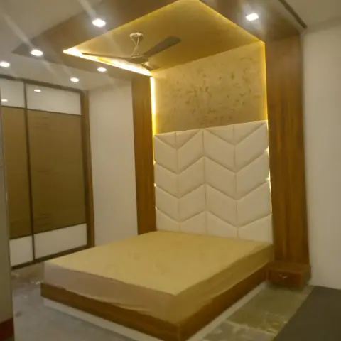 Newly finished bedroom with a chevron-quilted cream headboard set into a teak-laminate frame, a backlit gold textured panel above it