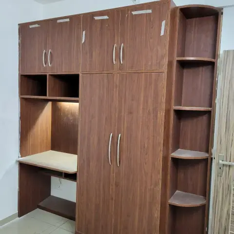 Newly installed wood-laminate wardrobe unit with an integrated study desk nook and open corner shelving, protective tape still on the doors.