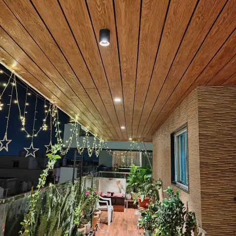 Night balcony under wood-grain PVC ceiling planks with a surface spotlight, star fairy lights strung along the glass railing and two rows of potted greenery
