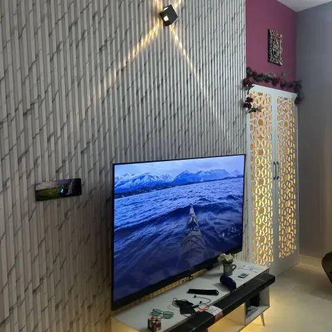 Night shot of a living-room TV wall in 3D fluted marble-look tile lit by a single up-down spot, the TV on a legged white console over a warm LED strip