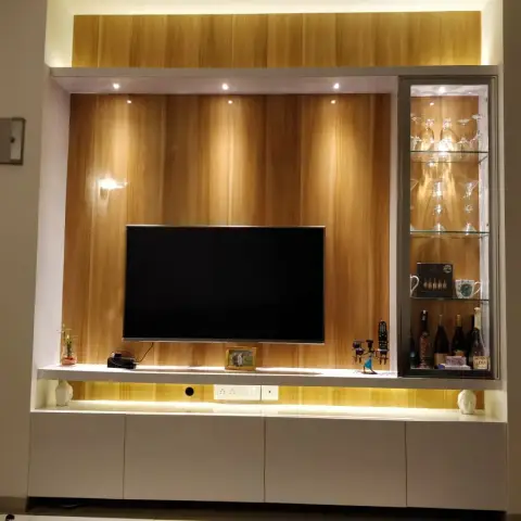 Night shot of a teak-veneer TV wall with spot-lit reveals, white gloss drawer bank below and a glass-fronted bar cabinet on the right holding upturned martini