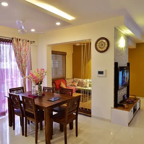 Open dining-and-living space with a wood dining table under pendant lighting, a TV wall in mustard paint, and a floor-seating swing nook beyond.