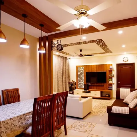 Open dining and living space with a wood dining table under copper pendant lights, a beamed wood ceiling, and a jaali-panelled TV unit wall visible beyond.