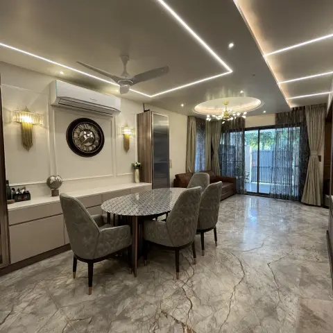 Open dining area with a round marble-veined table and tufted grey chairs under a multi-tier cove-lit false ceiling, opening onto a balcony.