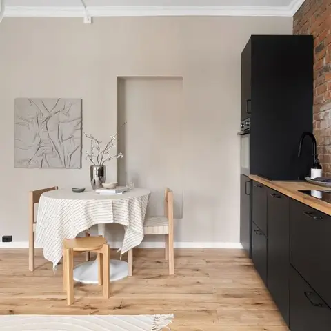 Open dining nook beside a matte-black galley kitchen with an exposed red-brick backsplash and wood countertop.